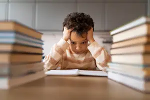 A child is sitting on a chair and studying in a room while holding their head in their hands.