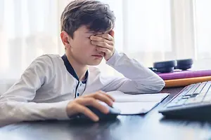 A young boy sitting at a desk rubbing his eyes while covering them with his hand.