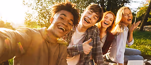 Four young people, including two women and two men, are sitting on the grass and posing for a selfie, smiling and laughing together