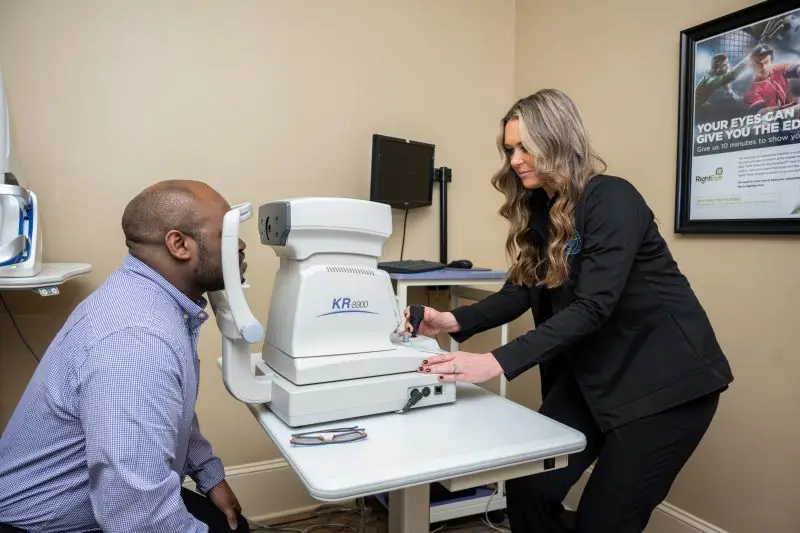 A man in a blue shirt is getting an eye exam from a woman in a black suit in a medical office with an eye exam machine on a table.