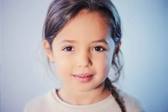 Portrait of a young girl with a smiling face and looking straight into the camera. She has braided hair and is wearing a white t-shirt.