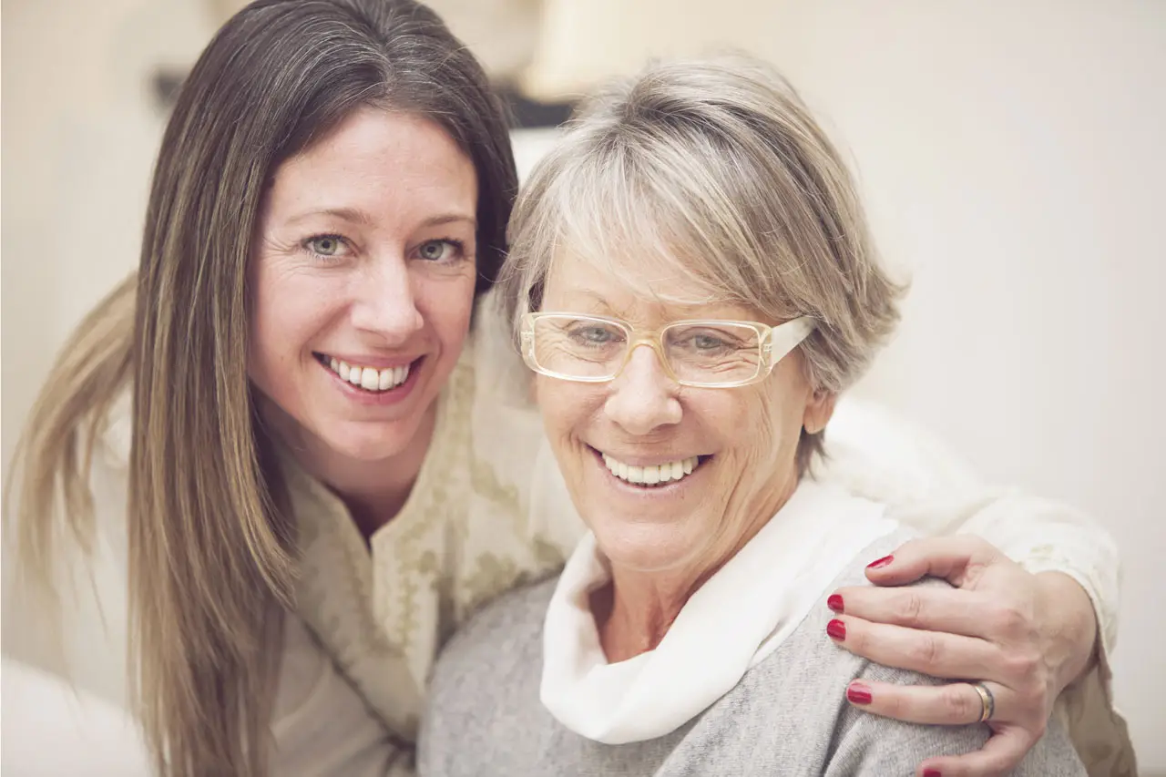 A woman with glasses and a white scarf is smiling and hugging an older woman in a white long-sleeve shirt.