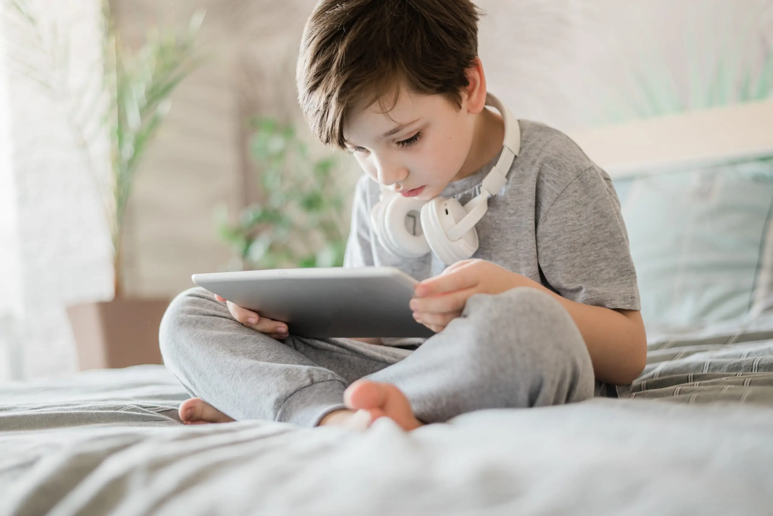 A boy is sitting on a bed and using a tablet with headphones on.