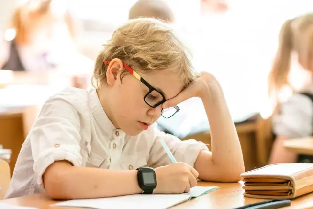 a young boy in a white shirt with glasses and a watch sitting at a desk writing in a notebook