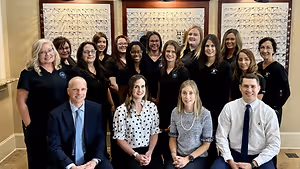 A group of people in a room, some are standing and some are sitting, all wearing black shirts, posing for a photo with a wall of eyeglasses in the background.