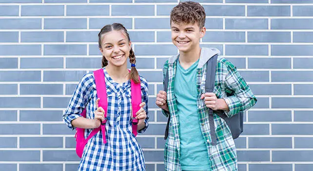 Two children, a girl and a boy, smiling and posing for a photo in front of a blue brick wall.
