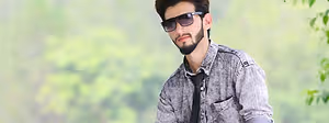 A man with a beard wearing sunglasses and a gray shirt is standing outside in a forest area with trees and plants in the background.