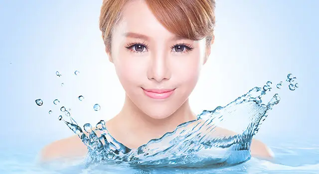 A woman with short hair and red hair is smiling with her teeth showing and her eyes closed. The water droplets are splashing around her face. She is probably in the swimming pool. The water droplets are sparkling and reflecting the light. The water droplets are splashing on her face, and the water droplets are splashing on the water.