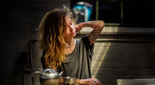 A woman sits on a chair in a kitchen, her eyes closed, a tattoo visible on her arm, a tea pot and cup on a table in front of her
