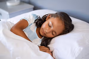 A young girl with long black hair sleeps peacefully in a bedroom, wearing orth-k contact lenses