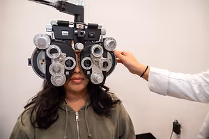 A woman is having her eyes examined by an optometrist wearing a green hoodie in a white wall background.