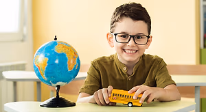 A young boy with glasses sits at a desk with a globe and a toy school bus.