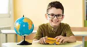 A young boy with glasses sits at a desk with a globe and a toy school bus.