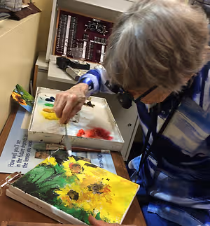 An elderly woman wearing glasses is painting a sunflower on a canvas in a room.