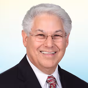 A man with white hair wearing a black suit, white shirt, and a red tie smiles for a photo.