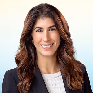 A smiling woman with brown wavy hair and brown eyes in a black blazer with white inner shirt standing in front of a blue and white background