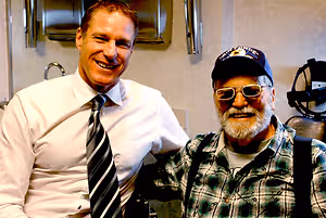 Two men pose for a photo in a room with a sink and mirror in the background. The man on the left is wearing a white shirt and tie, and the man on the right is wearing a plaid shirt and a baseball cap with the words 'Air Force' on it. Behind them, there is a mirror reflecting a sink and a towel dispenser, and on the right side, there is a black object resembling a helmet.
