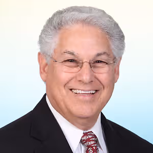 A smiling man with white hair and glasses wearing a black suit and a red tie is standing in front of a white background
