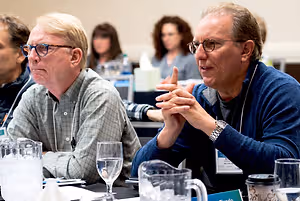 Two men wearing glasses sit at a table while a woman stands in the background in a classroom.