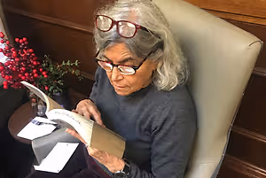 An older woman wearing glasses and a gray sweater is sitting in a chair reading a book with a red berry plant on a table next to her.