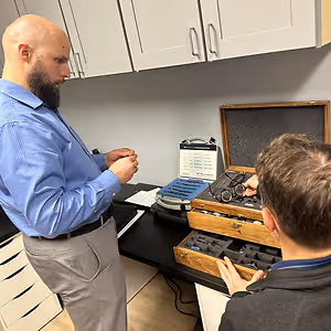 Two men are standing in a room, one wearing a blue shirt and the other a gray shirt, both looking at a wooden box with compartments on a black desk, likely discussing eyeglass fittings.