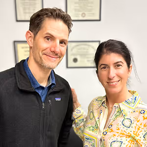 A man and a woman smiling at the camera with a white wall and certificates in the background