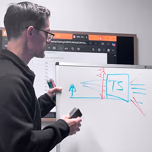 A man is writing on a whiteboard while standing in front of a computer screen