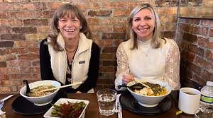 Two women sitting at a table in a restaurant, smiling and holding chopsticks, with bowls of food in front of them.