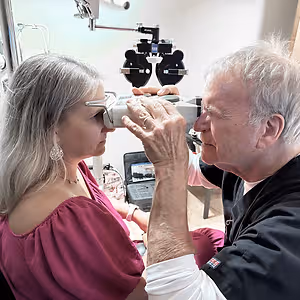 An elderly man examining the eyes of an elderly woman using a device in a room