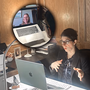 A woman is on a video call, a reflection of her is visible in the mirror of the laptop, and she is sitting at her desk with a computer and other items.