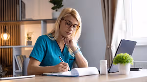 A woman in a blue scrub top is sitting at a desk and writing in a notebook.