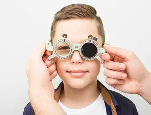 A young boy wearing green glasses with numbers and a cross symbol is looking at something in front of him while another person is holding his glasses in both hands.
