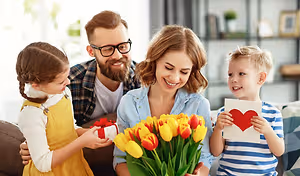 A family, including a woman, a man, and two children, is sitting on a couch, smiling and holding a bouquet of tulips and a gift