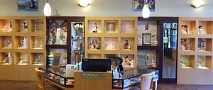 The interior of a shop displaying numerous framed glasses on wooden shelves with decorative elements and illuminated by ceiling lights, including a monitor on a desk with chairs in front.