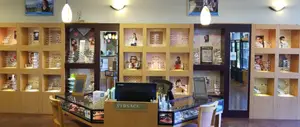 The interior of a shop displaying numerous framed glasses on wooden shelves with decorative elements and illuminated by ceiling lights, including a monitor on a desk with chairs in front.