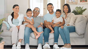 A family of five, including two young children, an elderly man, and two women, are sitting on a couch in a room, smiling and posing for a photo