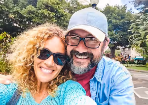 A man and a woman are smiling and posing for a picture in a park with trees and a building in the background.