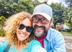 A man and a woman are smiling and posing for a picture in a park with trees and a building in the background.