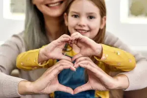 A woman and a young girl making a heart shape with their hands, smiling and posing for a picture in a room with a glass window and a white wall
