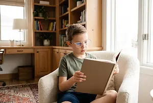 A boy with glasses is sitting in a chair and reading a book.