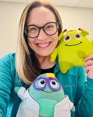 A woman wearing glasses is smiling and holding two plush toys, one with a green color and another with a purple and green color, and the woman is wearing a ring on her right hand and is dressed in a blue shirt. Behind her, there is a white wall.