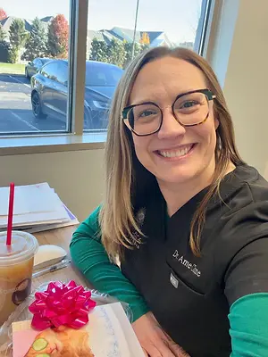 A smiling woman in a black uniform sits at a table with a cup of coffee, a pastry with a bow, and a book.