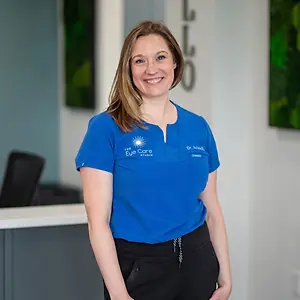 A smiling woman in blue shirt with The Eye Care Studio logo stands in front of a wall with green and white art.