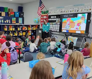 A woman teaching a group of children in a classroom using a monitor to show a cartoon.