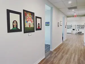 An art gallery with several paintings on the wall, a wooden floor, and a sign on the wall, along with a table and chair in the corner