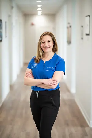 A smiling woman wearing a blue shirt and black pants is standing in a hallway with her arms crossed