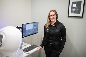 A woman in a black jacket and glasses stands in a room next to a desk with a computer monitor and keyboard, and a large white machine resembling a helmet with a blue light. The room has a white wall with a picture frame and a blue light on the wall.