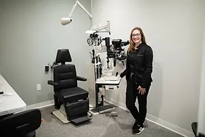 A woman with glasses stands in a room with a desk and an eye examination chair