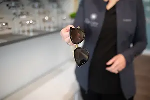 A person is holding a pair of sunglasses in their hand, and they are standing inside a shop, probably a shop that sells sunglasses. The person is wearing a black shirt and a blazer with a logo on the left side. The shop has a white wall, and the glass display has several objects on it, such as bottles and glasses. There is also a blurry view of a potted plant in the distance.