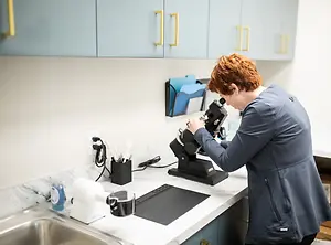 A woman wearing a lab coat and a blue blazer is looking into a microscope in a lab room